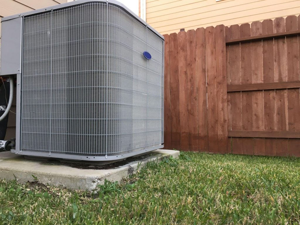 An outdoor AC unit in a backyard with grass and a brown fence